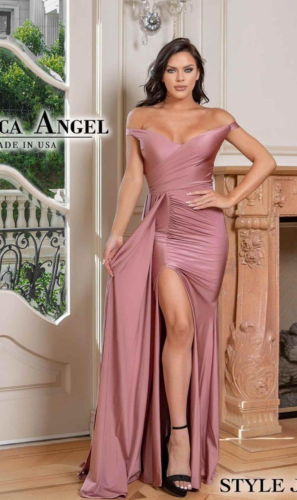 A woman poses indoors by a decorative fireplace, wearing the Jessica Angel 776 Side-Drape Long Prom Dress—an elegant mauve off-the-shoulder gown with a thigh-high slit.